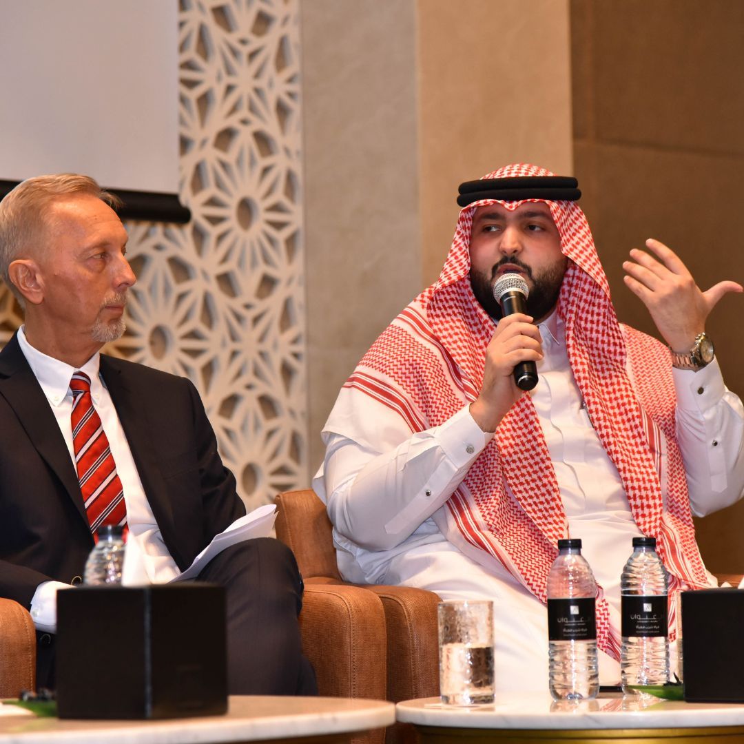 A delegate in a red-and-white shemagh speaks into a microphone during a seated panel discussion.